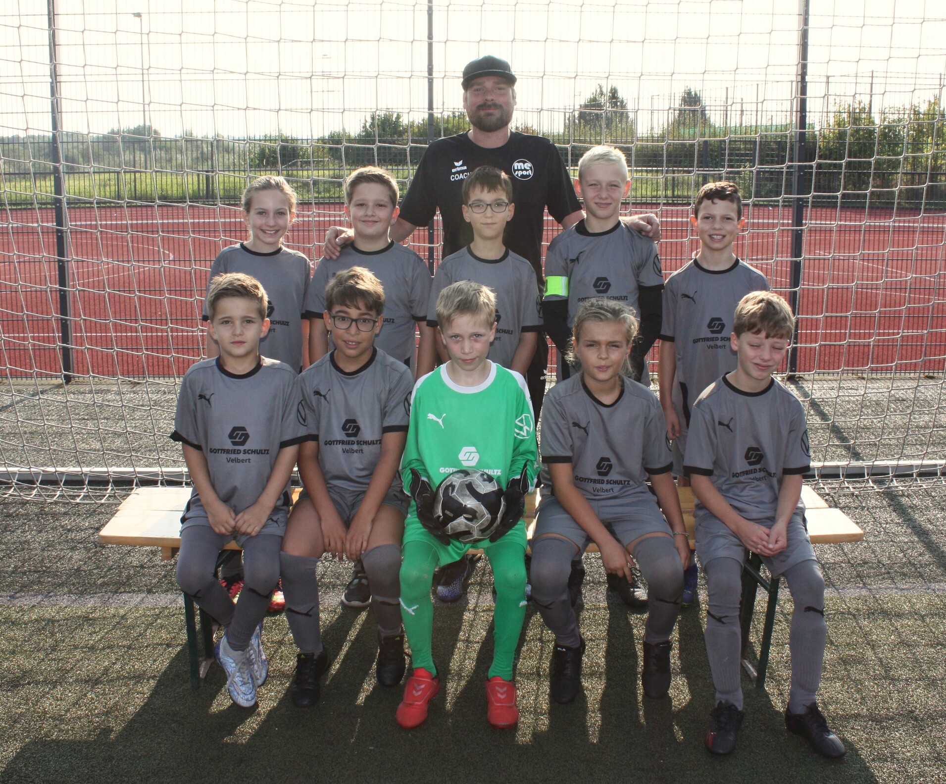 Fußballmannschaft von Kindern in grauen Trikots und Trainer, auf dem Sportplatz vor einem Tor, lächelnd posierend.