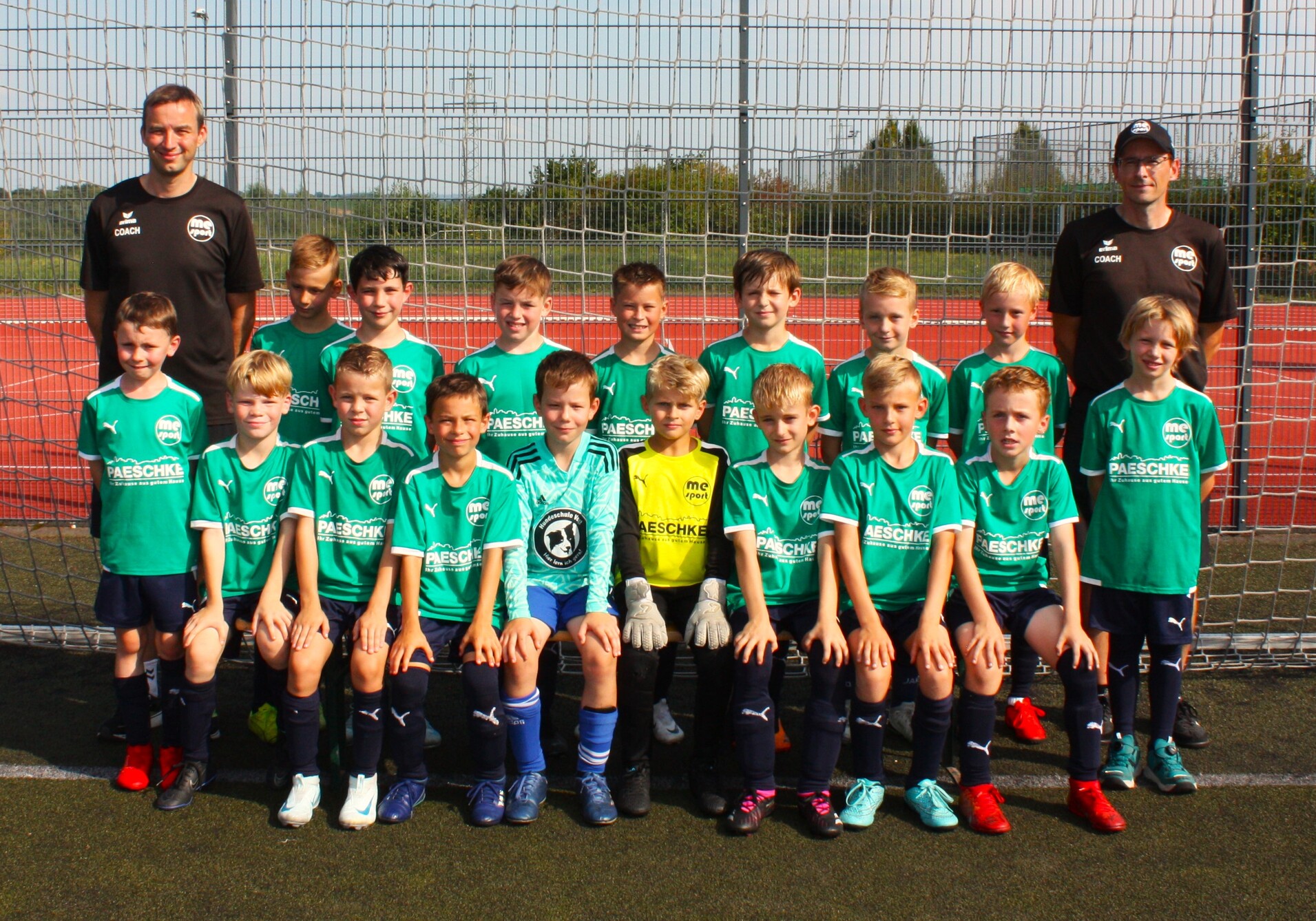 Gruppenfoto einer Jugendfußballmannschaft mit Trainer, alle tragen grüne Trikots und stehen vor einem Fußballtor.
