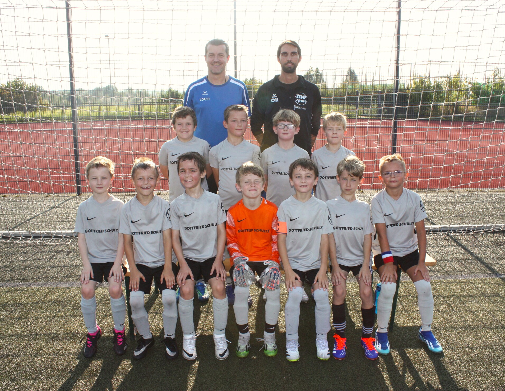 Kinderfußballmannschaft in grauen Trikots posiert vor einem Fußballtor auf einem Kunstrasenplatz.