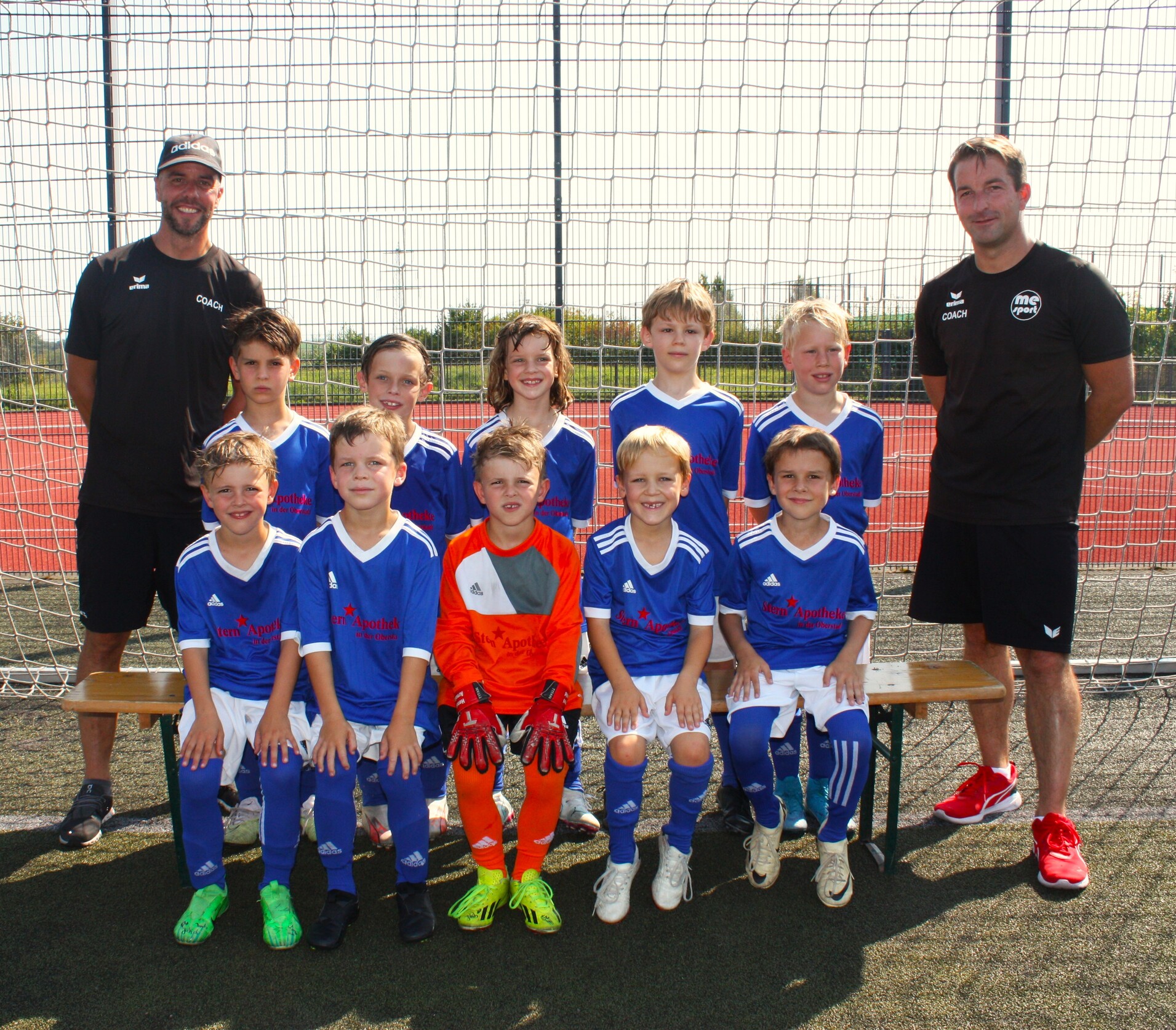 Gruppenshot einer Jugendfußballmannschaft in blauen Trikots, zwei Trainer stehen hinter den Spielern vor einem Tor.