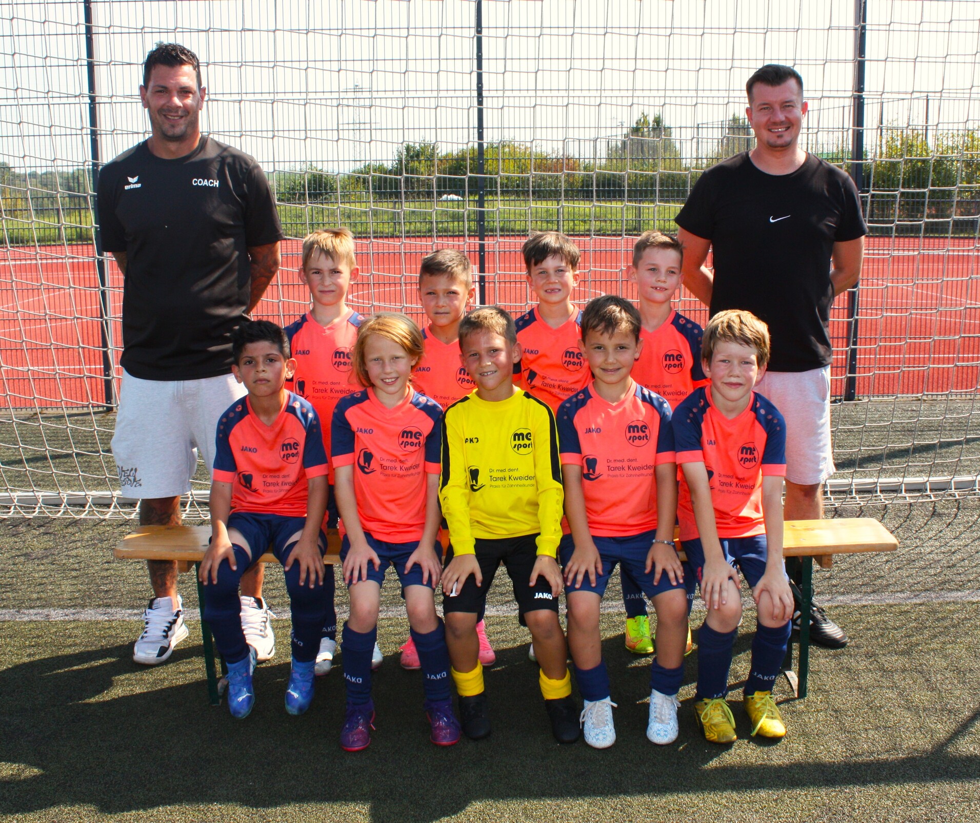 Kinderfußballmannschaft posiert mit Trainern vor einem Fußballtor auf einem Kunstrasenplatz.