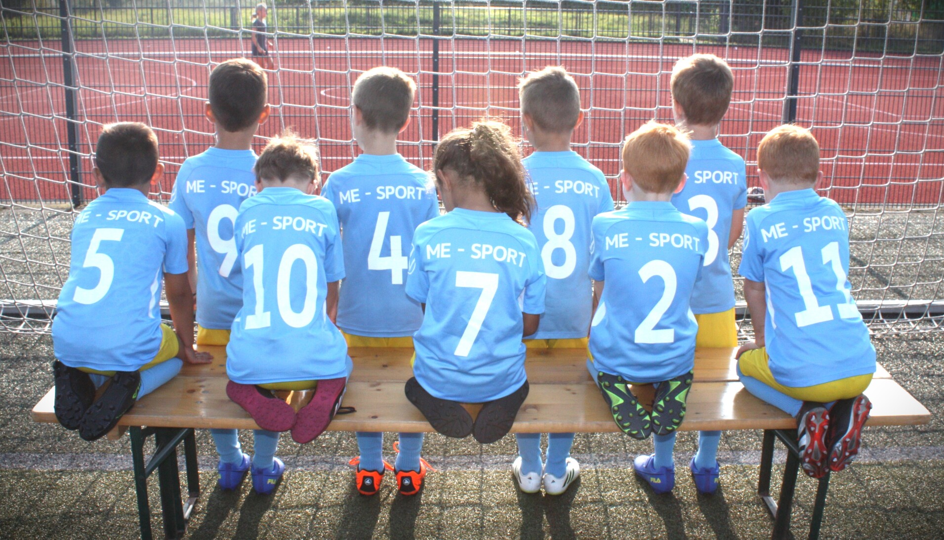 Gruppe von Kindern in blauen Trikots mit Nummern, sitzend auf einer Bank vor einem Fußballtor.