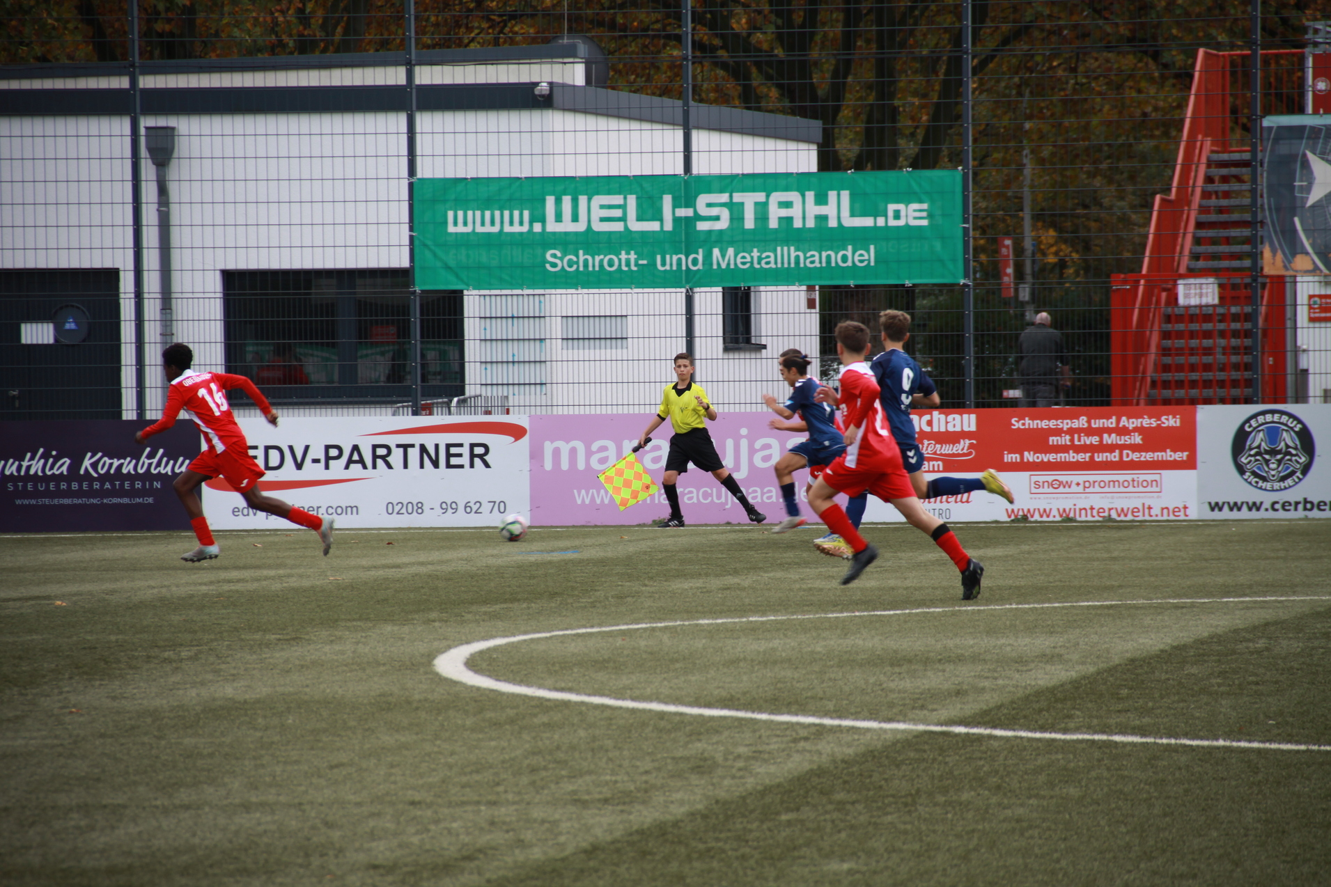 Junger Fußballspieler in roter Uniform sprintet während eines Spiels über den Kunstrasen, Schiedsrichter beobachtet.