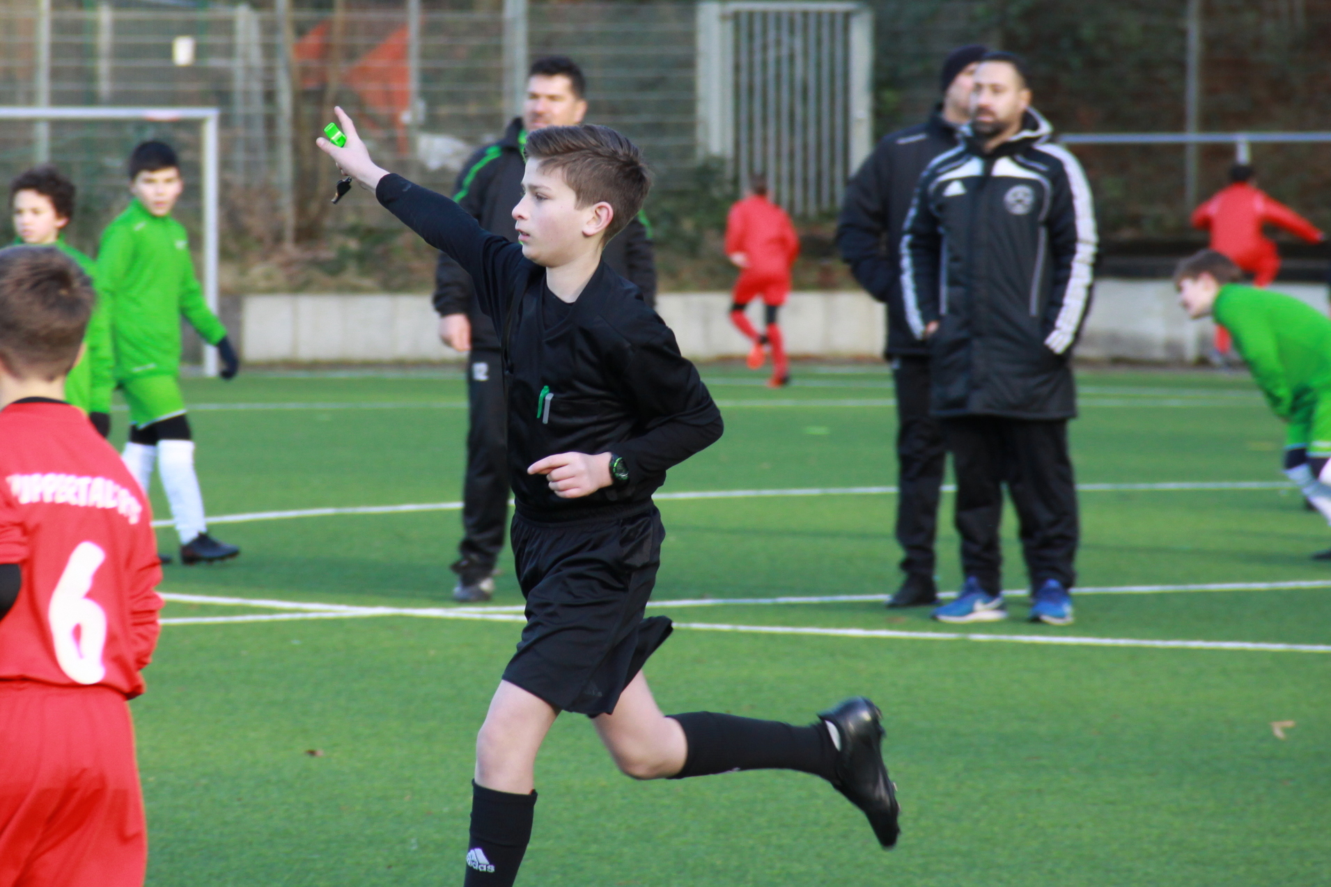 Ein junger Schiedsrichter signalisiert auf dem Fu&szlig;ballfeld, w&auml;hrend Spieler in roten und gr&uuml;nen Trikots im Hintergrund aktiv sind.