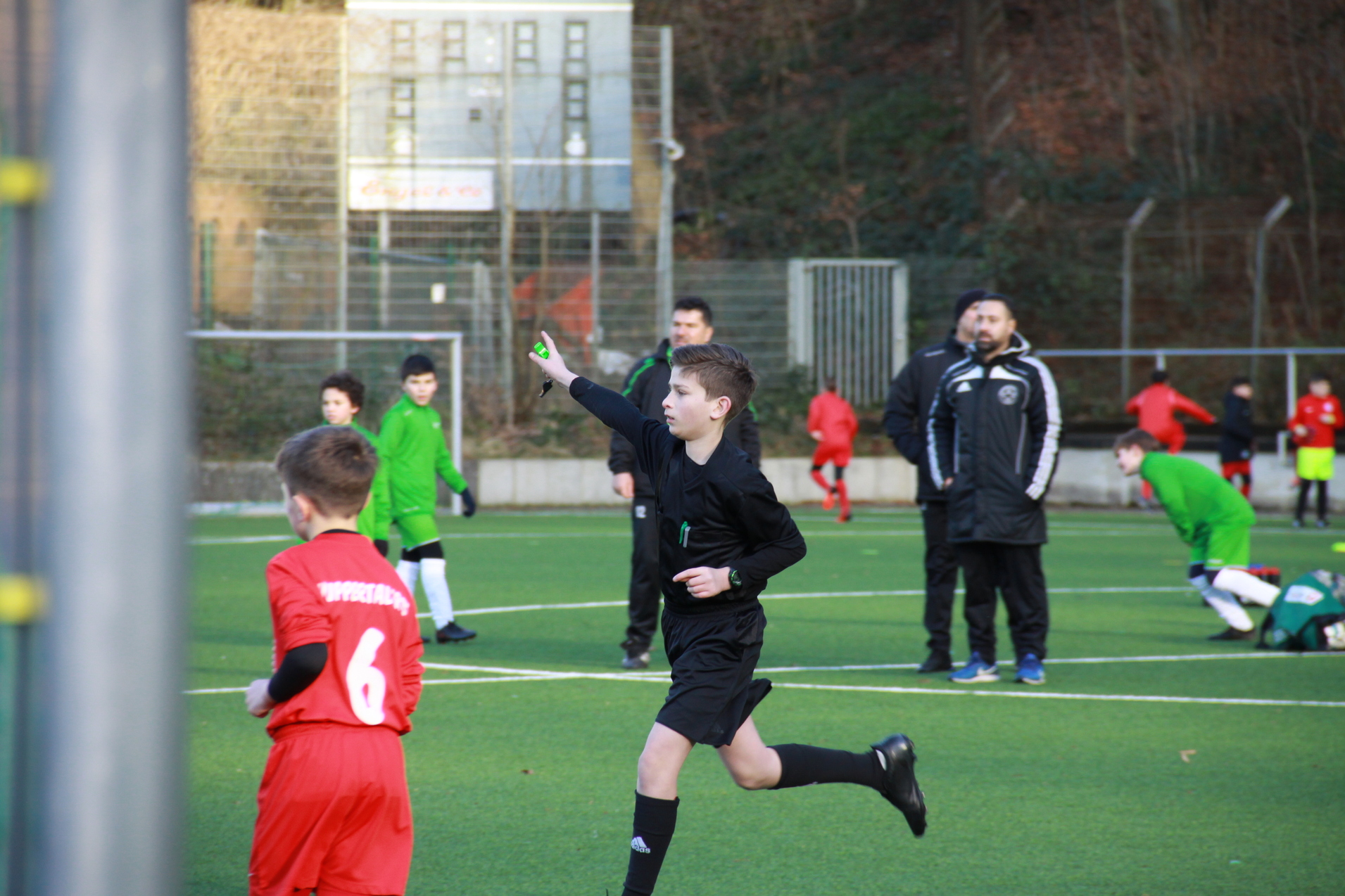 Ein junger Schiedsrichter signalisiert auf dem Fußballfeld, während Spieler in roten und grünen Trikots im Hintergrund aktiv sind.