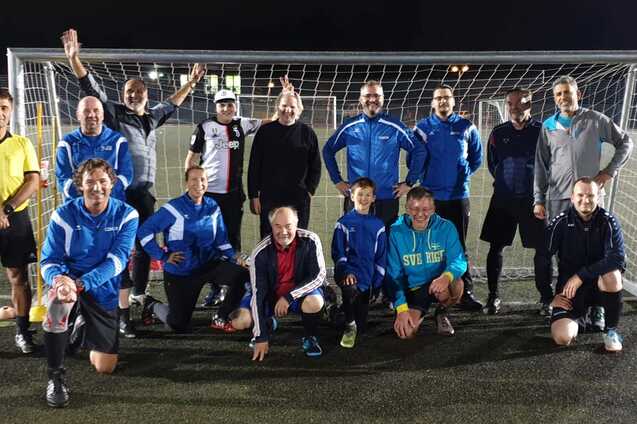 Gruppe von 15 Erwachsenen und einem Kind posieren in Sportkleidung vor einem Fußballtor bei Nacht.