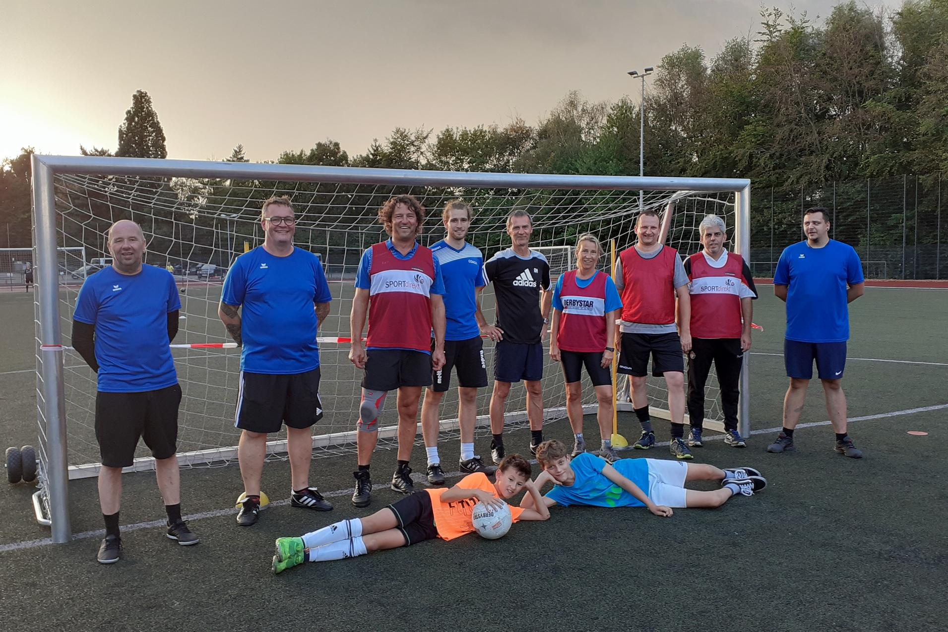 Gruppenshooting von Fußballspielenden Erwachsenen und Kindern vor einem Tor auf einem Kunstrasenplatz.