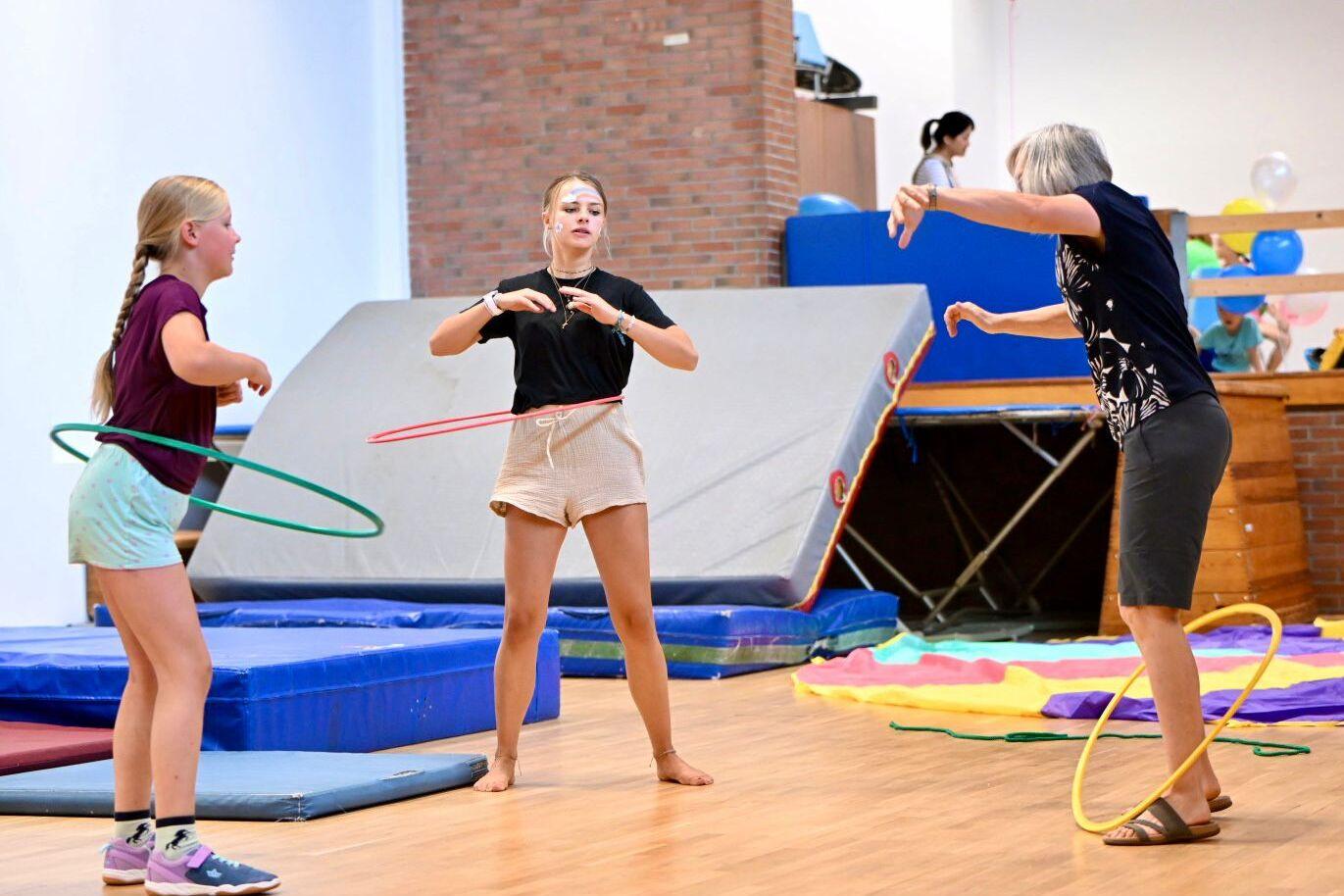 Drei Personen in einer Turnhalle jonglieren mit Hula-Hoops auf einer Holzbodenfl&auml;che umgeben von Turnmatten.