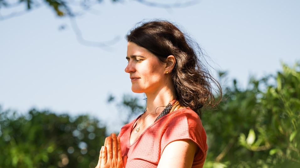 Frau in rotem Shirt im Baum-Yoga, steht auf einem Bein und h&auml;lt die H&auml;nde in Gebetshaltung vor der Brust.