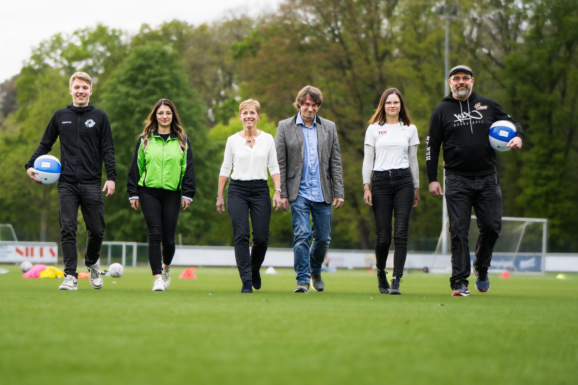 Sechs Personen gehen auf einem Fu&szlig;ballplatz, zwei halten Volleyball, umgeben von Trainingsmaterialien und gr&uuml;ner Natur.