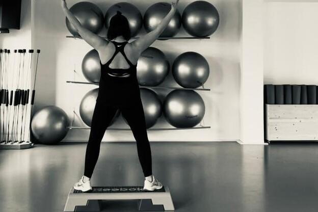 Frau mit Sportoberteil steht auf einer Step-Box und hebt die Arme in einem modernen Fitnessstudio mit Gymnastikbällen.