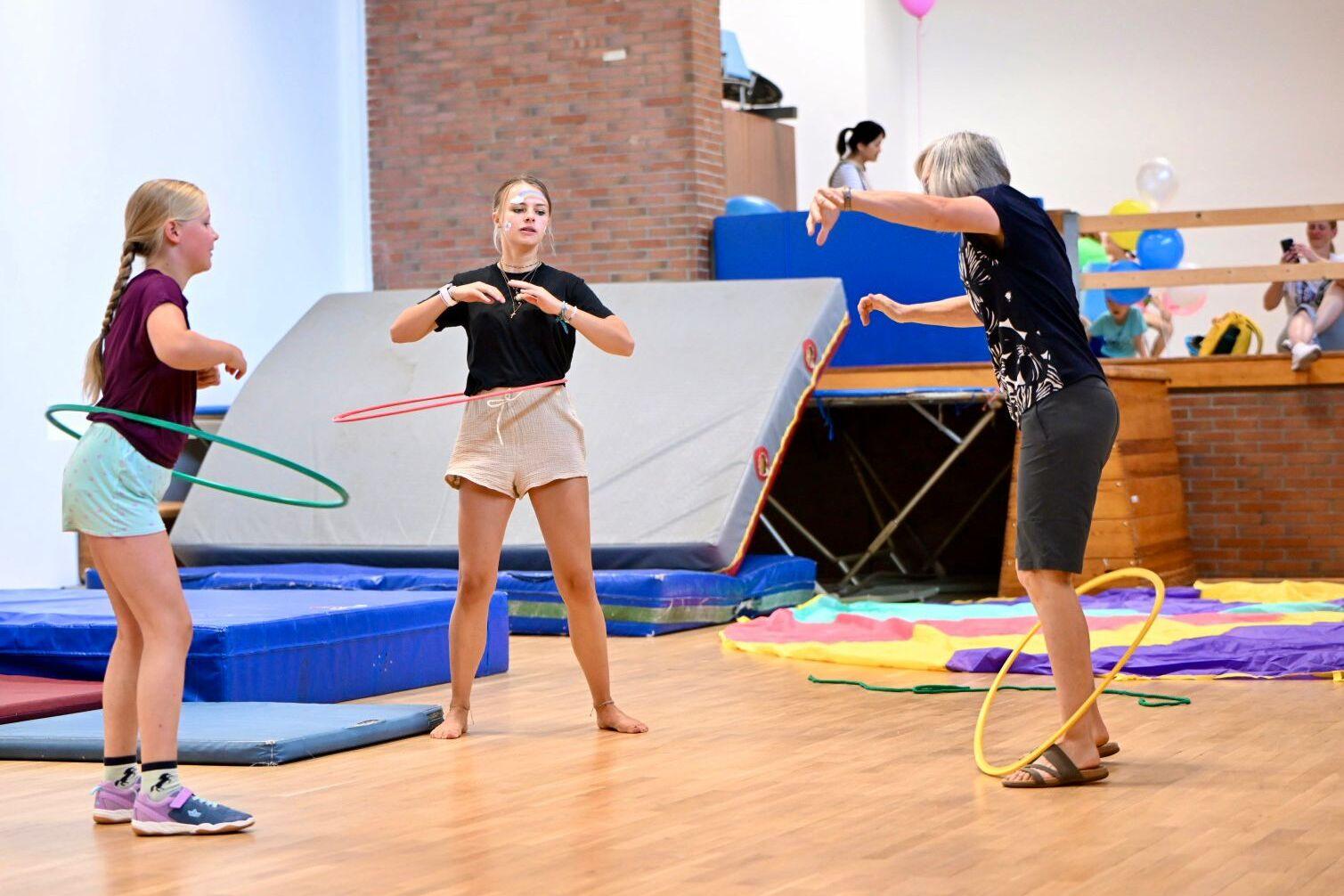 Drei Personen in einer Turnhalle jonglieren mit Hula-Hoops auf einer Holzbodenfläche umgeben von Turnmatten.
