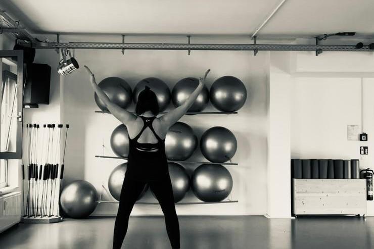 Frau mit Sportoberteil steht auf einer Step-Box und hebt die Arme in einem modernen Fitnessstudio mit Gymnastikbällen.