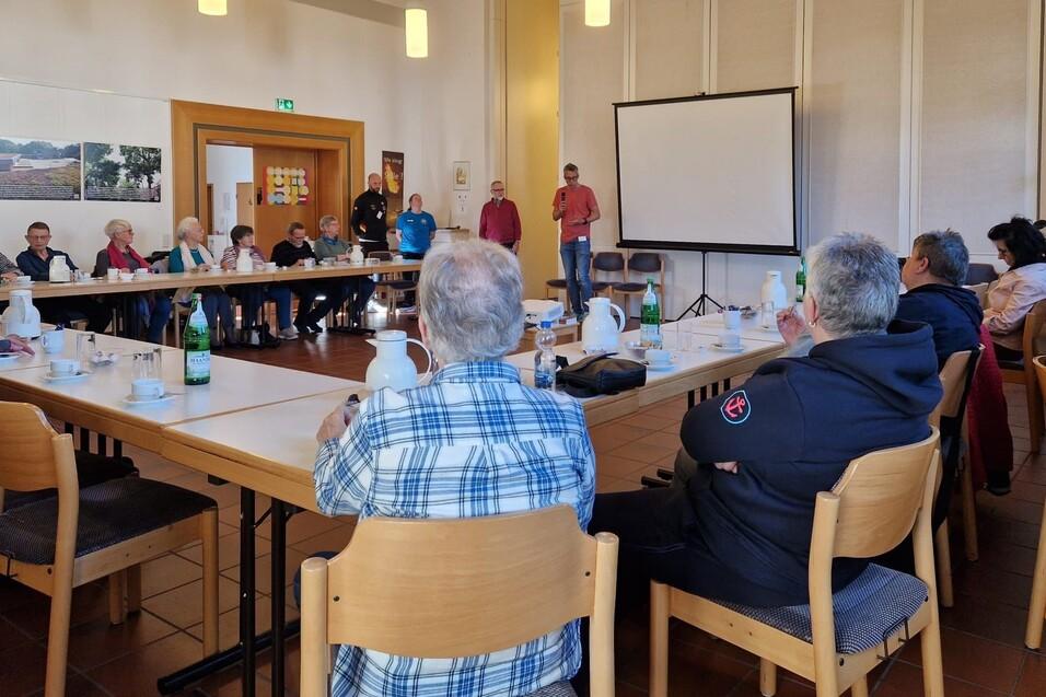 Gruppe von Menschen sitzt an Tischen in einem Raum, während ein Redner vor einer Leinwand spricht.