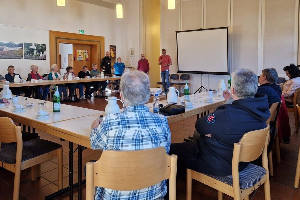 Gruppe von Menschen sitzt an Tischen in einem Raum, während ein Redner vor einer Leinwand spricht.