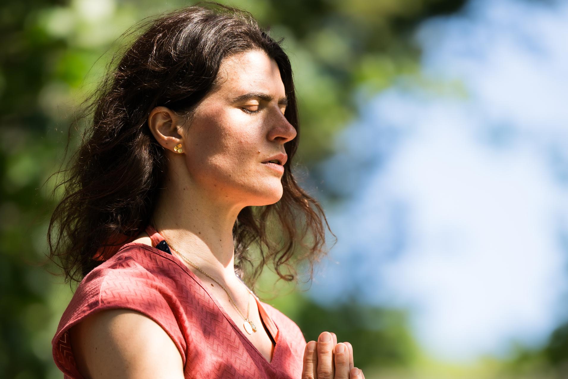 Frau sitzt im Freien, meditierend mit gefalteten H&auml;nden, umgeben von gr&uuml;ner Natur und sanftem Licht.
