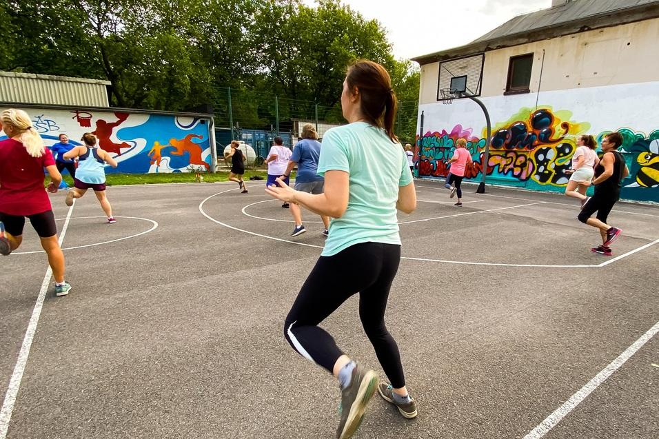 Gruppe von Frauen joggt auf einem Sportplatz mit buntem Graffiti-Hintergrund und Basketballkorb.