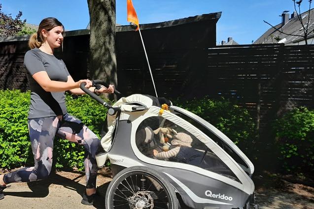 Frau joggt mit einem Kinderanh&auml;nger auf einem Weg, umgeben von gr&uuml;nen Pflanzen und blauem Himmel.