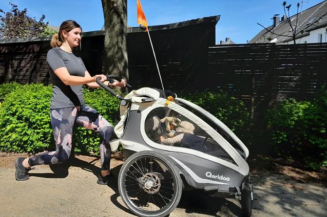 Frau joggt mit einem Kinderanhänger auf einem Weg, umgeben von grünen Pflanzen und blauem Himmel.