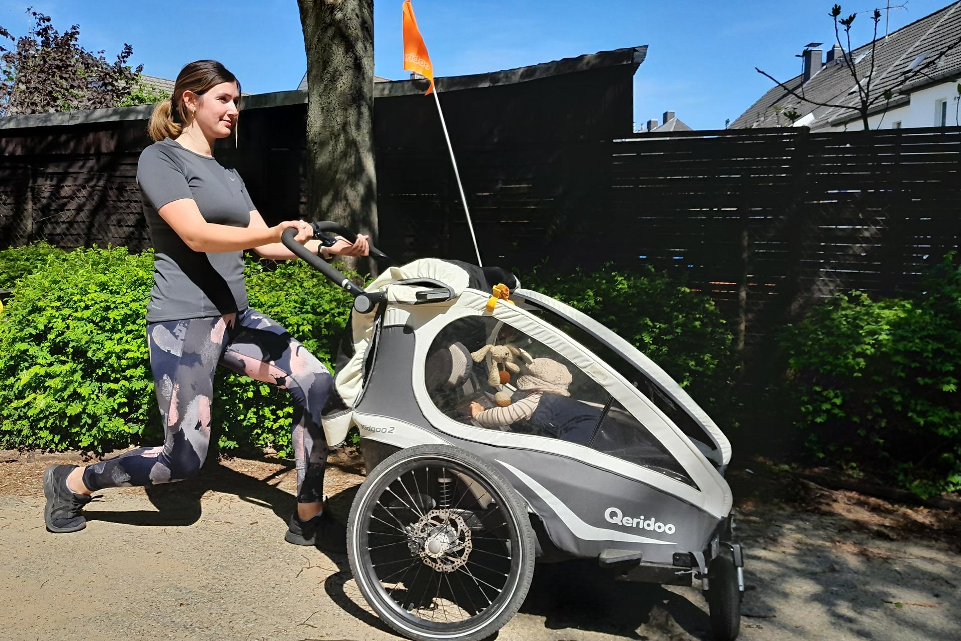 Frau joggt mit einem Kinderanhänger auf einem Weg, umgeben von grünen Pflanzen und blauem Himmel.