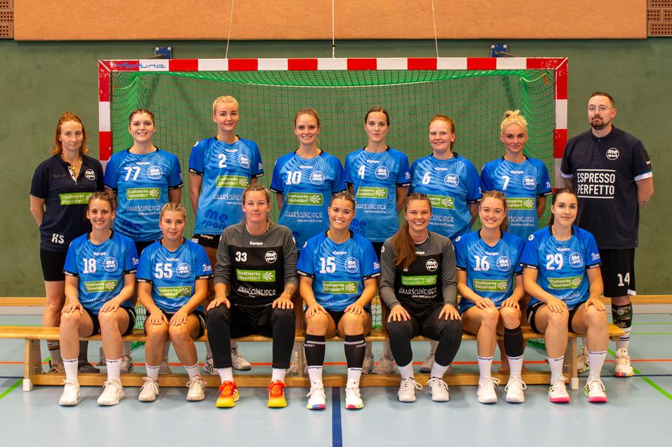 Gruppenfoto eines Handballteams mit 14 Spielerinnen und Trainern in blauen Trikots vor einem Handballtor.