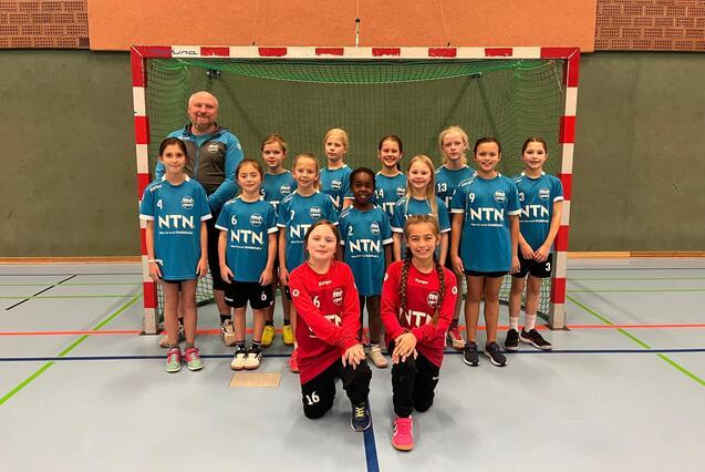 Mannschaftsbild einer Handballjugend mit Trainer, alle in blauen Trikots, vor einem Handballtor in einer Sporthalle.