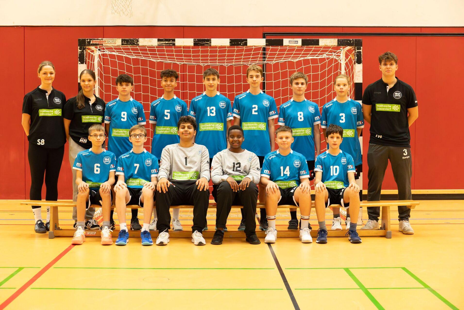 Mannschaftsfoto einer Handballjugend mit Spielern in blauen Trikots und zwei Trainerinnen vor einem Tor.