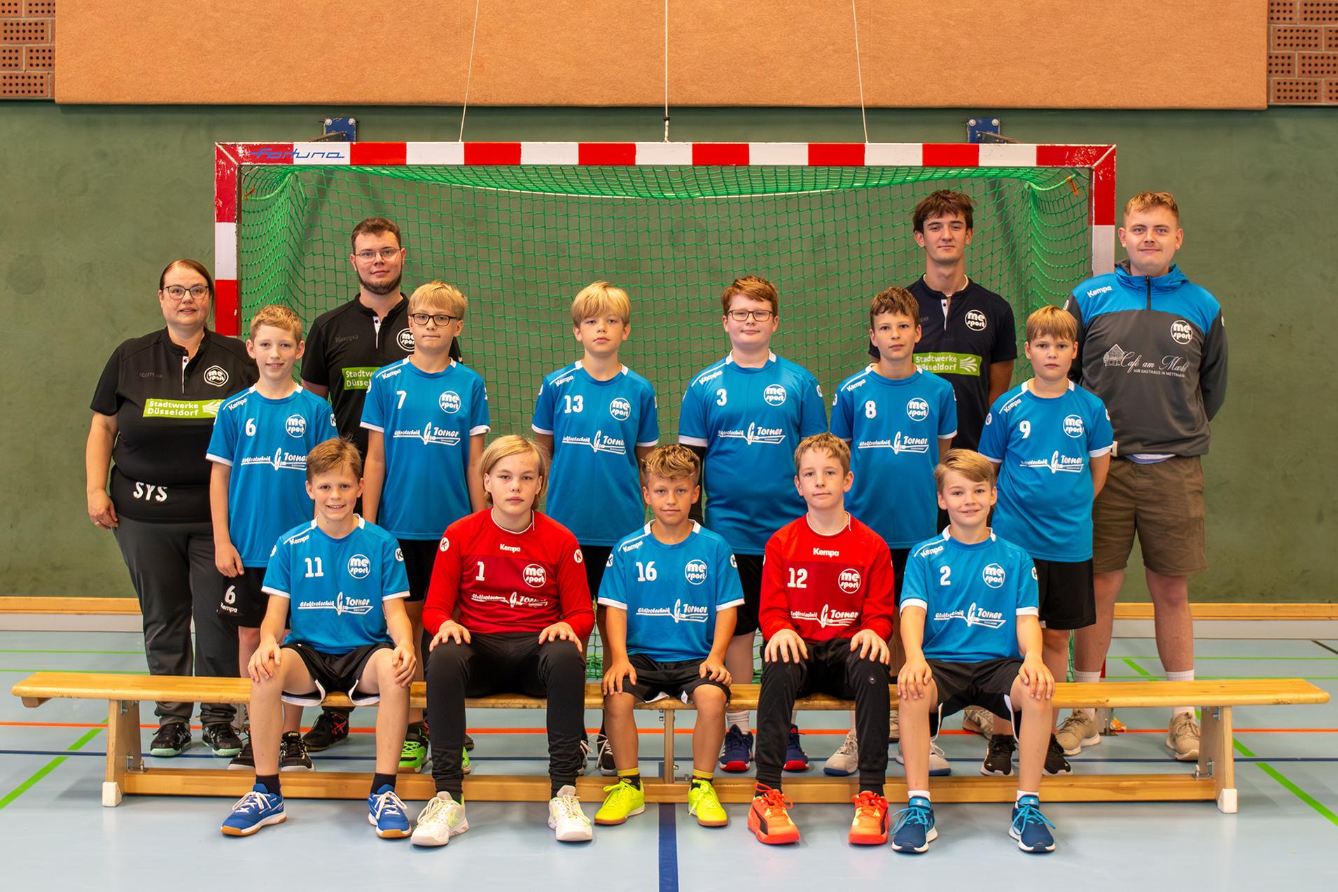 Gruppenfoto einer Handballmannschaft mit Trainer, in blauen und roten Trikots, vor einem Tor auf einem Sportfeld.