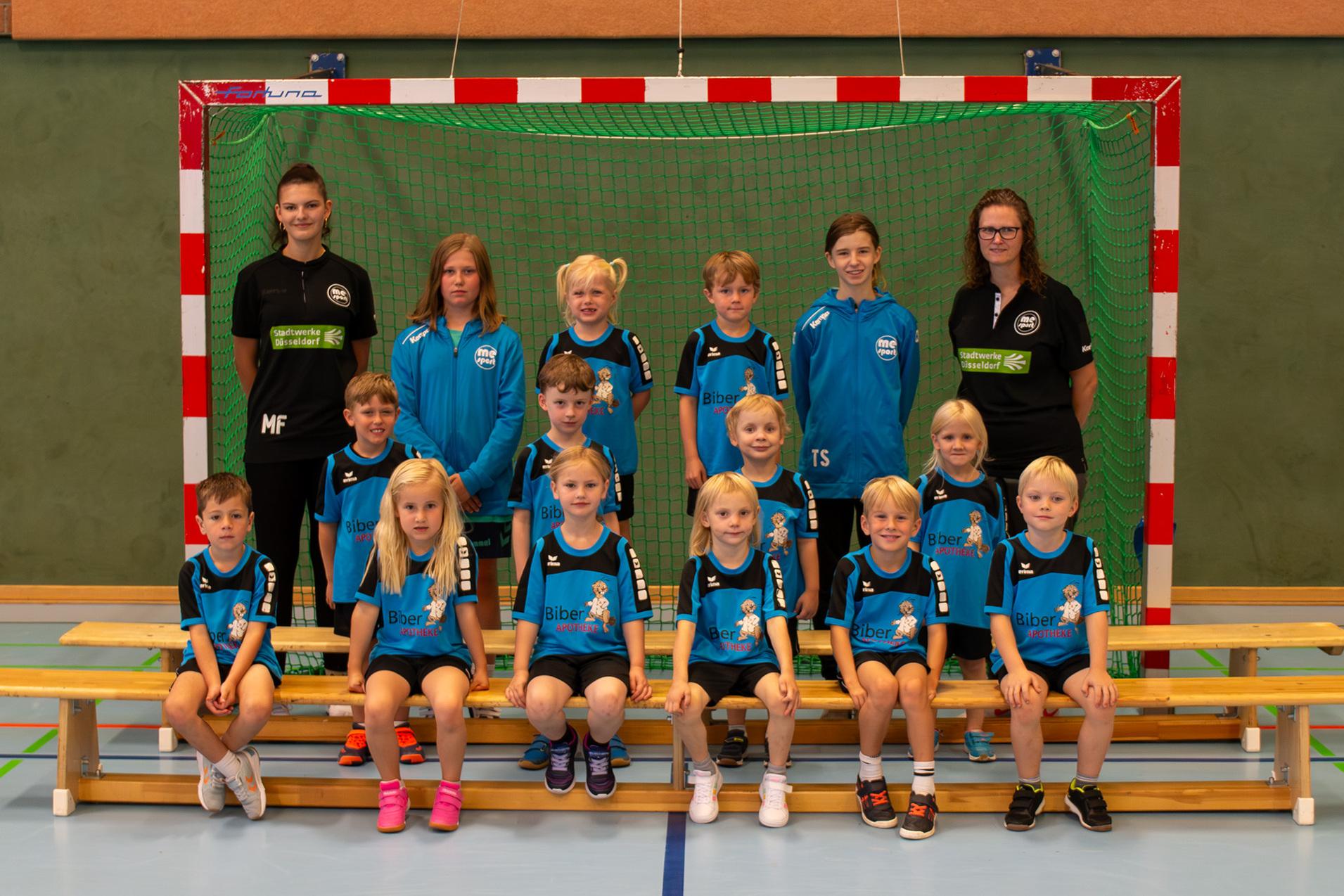 Gruppenfoto einer Jugendmannschaft mit Trainern in blauen Trikots vor einem Handballtor in einer Sporthalle.