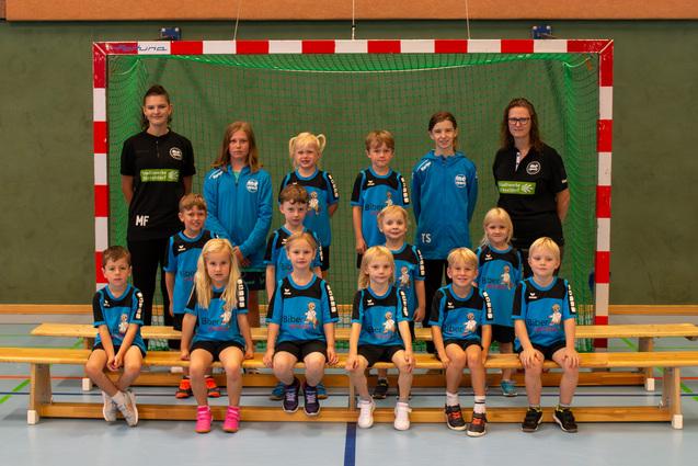 Gruppenfoto einer Jugendmannschaft mit Trainern in blauen Trikots vor einem Handballtor in einer Sporthalle.