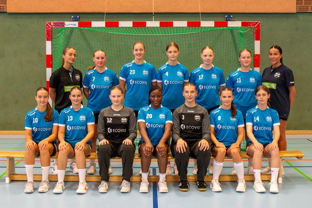 Mannschaftsfoto einer Handballmannschaft mit 14 Spielerinnen und Trainerin vor einem Tor in der Sporthalle.