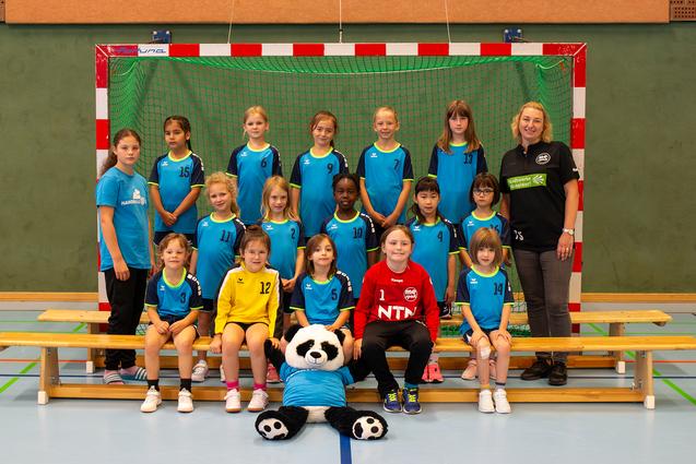 Mannschaftsfoto einer Handballjugend mit 14 Mädchen und einer Trainerin, sitzend vor einem Handballtor in einer Sporthalle.