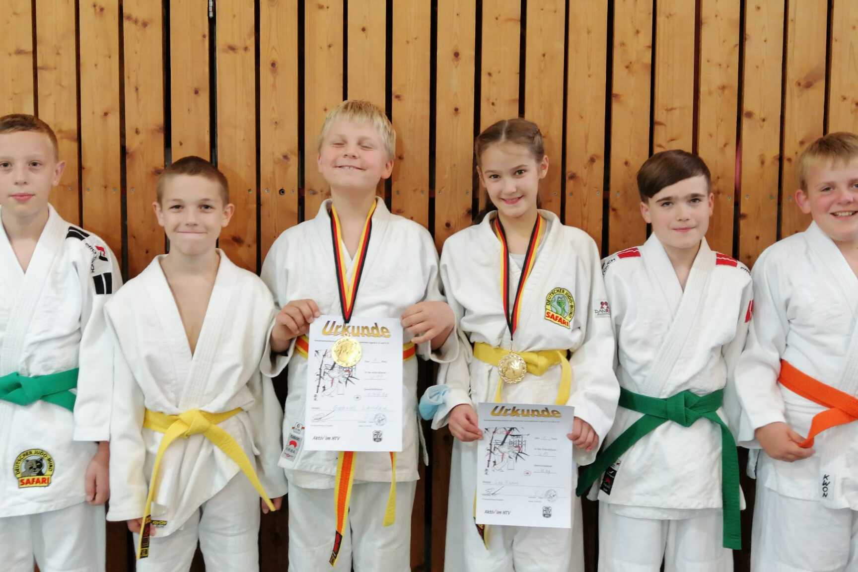Sechs Kinder in Judoanz&uuml;gen stehen vor einer Holzwand und halten Urkunden, einige mit Medaillen um den Hals.