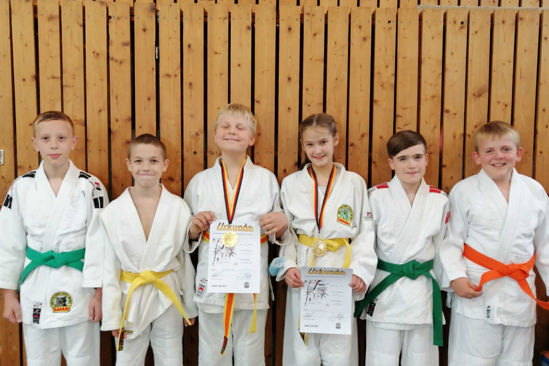 Sechs Kinder in Judoanzügen stehen vor einer Holzwand und halten Urkunden, einige mit Medaillen um den Hals.