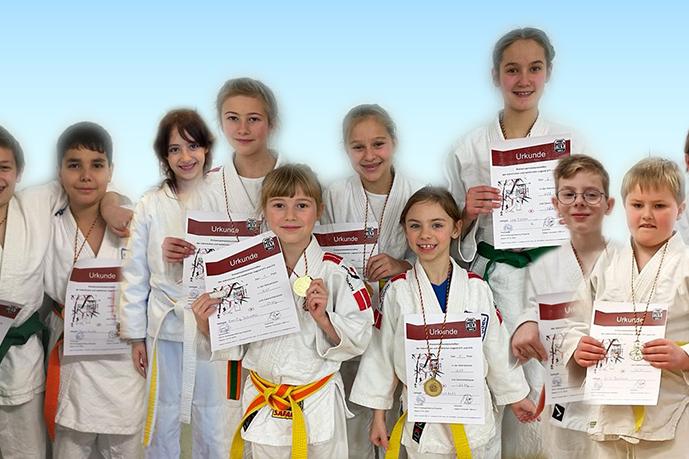 Gruppe von zehn Kindern in Judoanzügen, die Urkunden und Medaillen stolz halten. Hintergrund ist ein heller Farbverlauf.