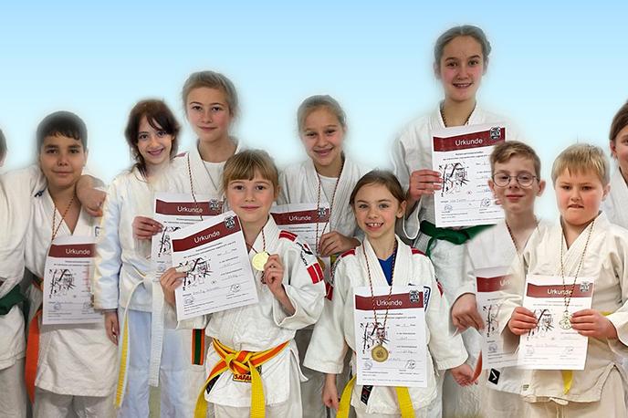 Gruppe von zehn Kindern in Judoanz&uuml;gen, die Urkunden und Medaillen stolz halten. Hintergrund ist ein heller Farbverlauf.