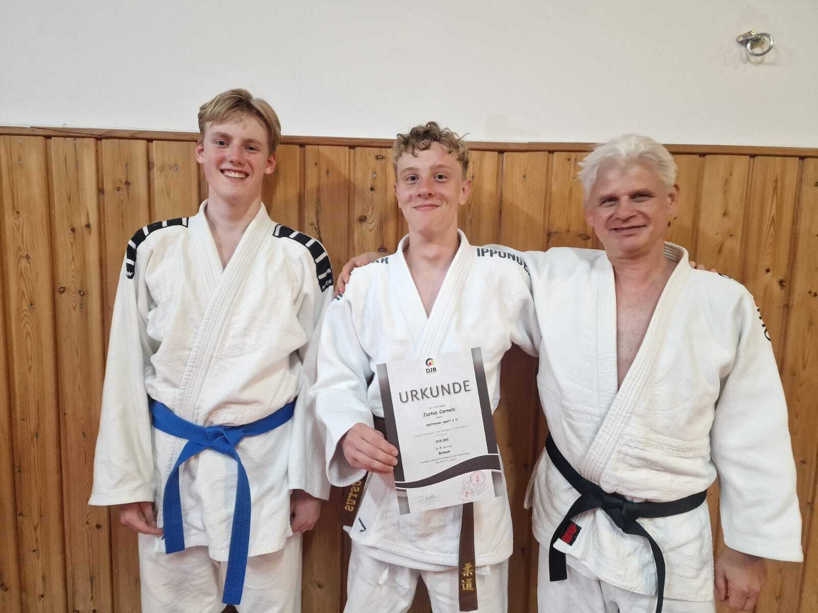 Drei Judoka in weißen Gi, einer hält eine Urkunde, stehen vor einer Holzwand in einem Dojo.
