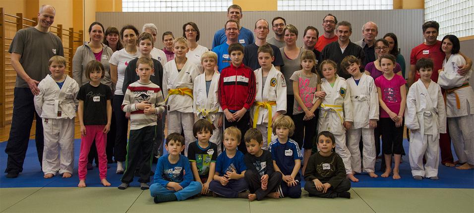 Gruppenfoto von Erwachsenen und Kindern in Judokleidung auf einer Mattenfläche in einem Sportstudio.