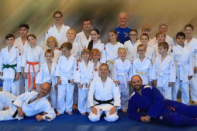 Gruppe von Judokas in weißen und blauen Judogis auf einer Mattenfläche, um einen Trainer und Teilnehmer zu versammeln.