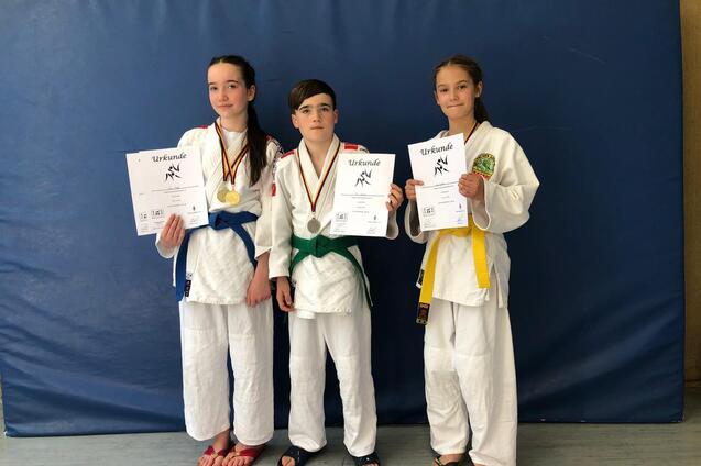 Drei Jugendliche in Karate-Gewändern posieren mit Urkunden und Medaillen vor einer blauen Wand.