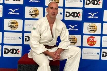 Mann im Judoanzug sitzt auf einer roten Bank vor Werbebanner der Deutschen Meisterschaften in Bad Homburg 2021.