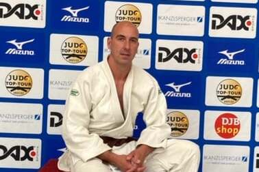 Mann im Judoanzug sitzt auf einer roten Bank vor Werbebanner der Deutschen Meisterschaften in Bad Homburg 2021.
