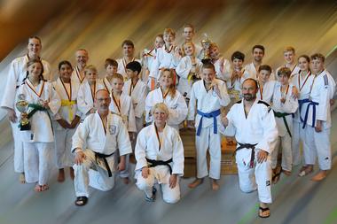 Gruppe von Judo-Trainierenden in weißen Judogis, einige halten Pokale und Medaillen, in einem Sportstudio.