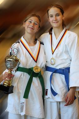 Zwei junge Judokas in weißen Judogi halten einen Pokal und Medaillen, lächeln freundlich in die Kamera.