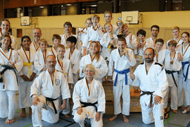 Gruppe von Judokas in Weiß und Farben, posierend nach einem Wettkampf mit Medaillen und Trophäen in einer Sporthalle.