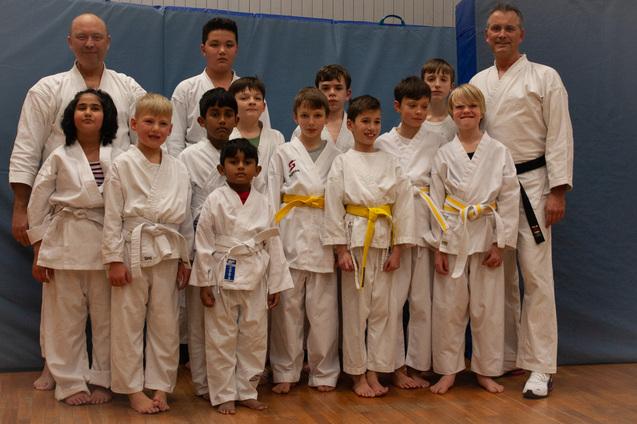 Gruppe von Jugendlichen im Karateanzug mit verschiedenen Gürtelfarben steht vor einer blauen Wand.