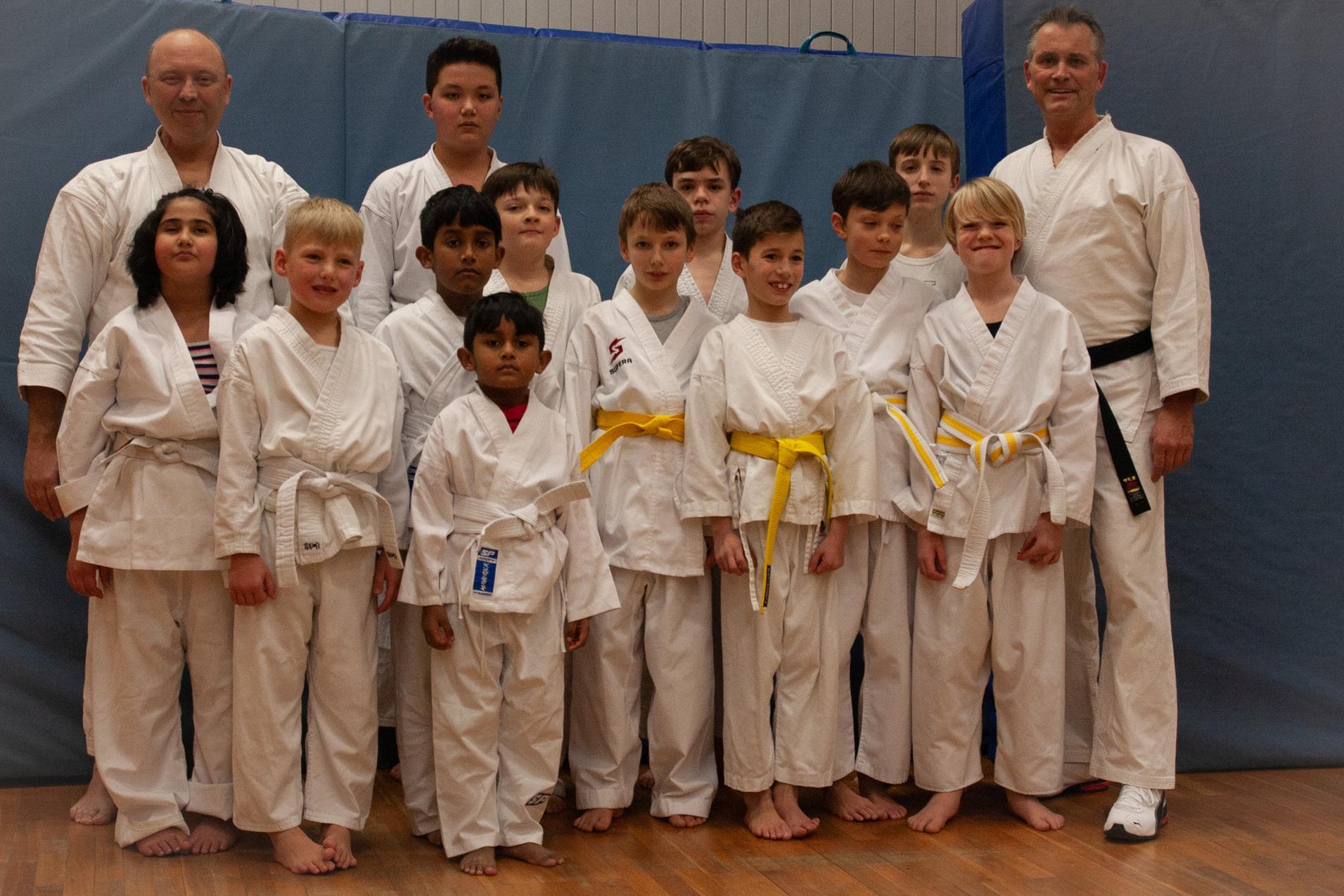 Gruppe von Jugendlichen im Karateanzug mit verschiedenen Gürtelfarben steht vor einer blauen Wand.