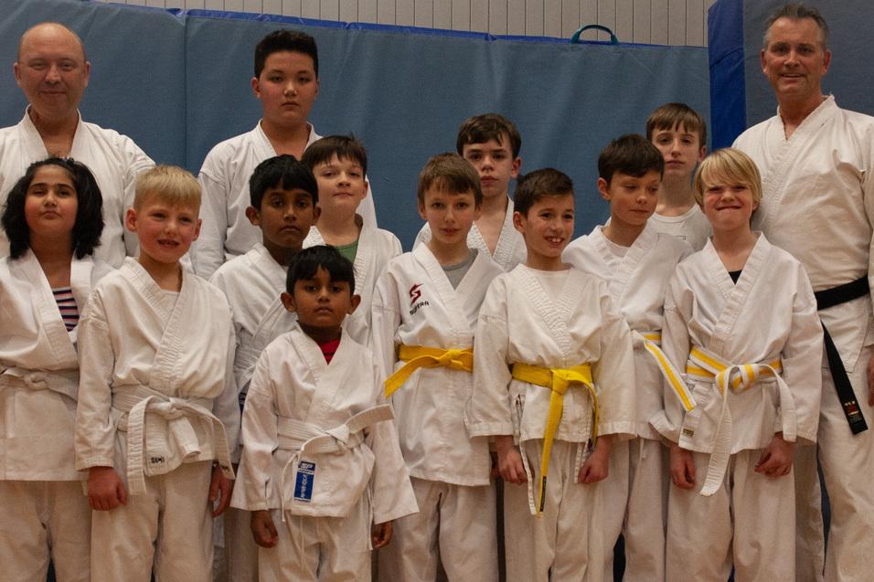 Gruppe von Jugendlichen im Karateanzug mit verschiedenen G&uuml;rtelfarben steht vor einer blauen Wand.