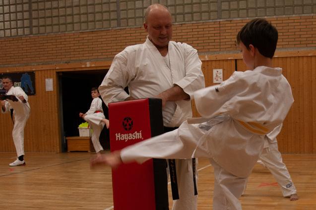 Junge in Karate-Duniform tritt gegen einen gepolsterten Schlagblock, w&auml;hrend ein Trainer ihn anleitet.