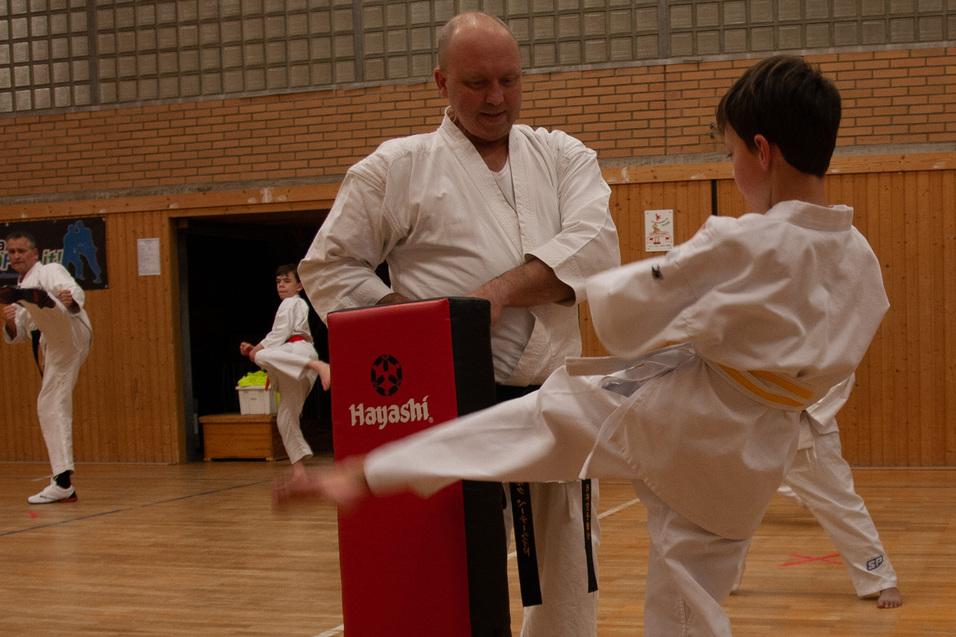 Junge in Karate-Duniform tritt gegen einen gepolsterten Schlagblock, w&auml;hrend ein Trainer ihn anleitet.